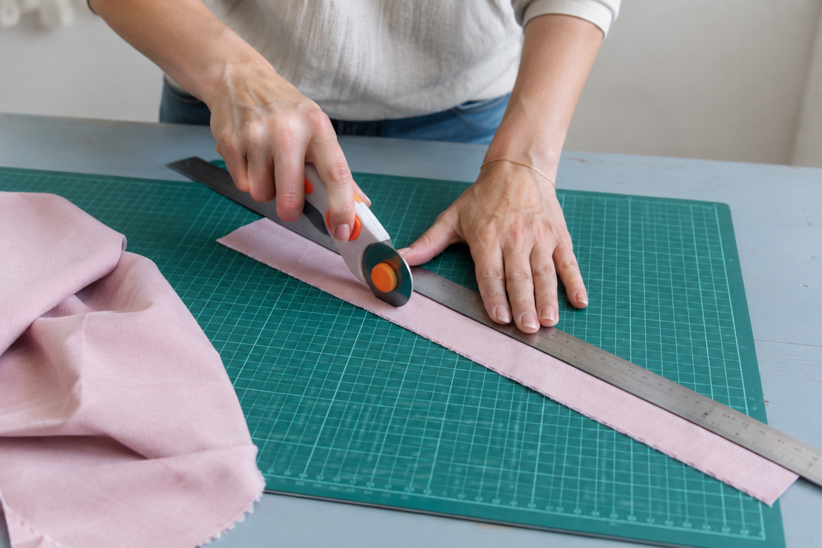 Mains de femme coupant une bande de tissu rose poudré au cutter rotatif sur une planche de découpe verte 45x30 cm